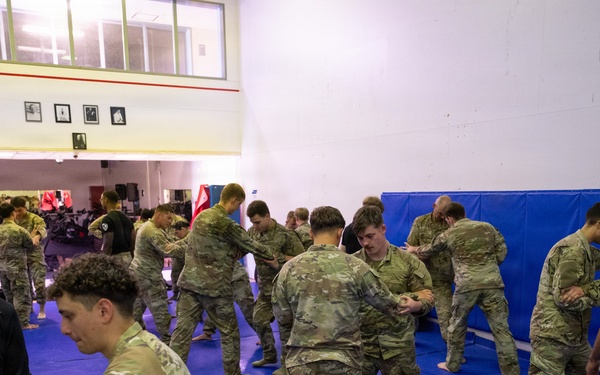 A group of military personnel in camouflage uniforms are conducting combatives or hand-to-hand combat training on blue gymnasium mats. The soldiers are paired up and practicing grappling or defensive techniques in what appears to be an indoor training facility. The setting includes wall-mounted crash pads and an adjacent weight room visible in the background.