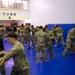A group of military personnel in camouflage uniforms are conducting combatives or hand-to-hand combat training on blue gymnasium mats. The soldiers are paired up and practicing grappling or defensive techniques in what appears to be an indoor training facility. The setting includes wall-mounted crash pads and an adjacent weight room visible in the background.