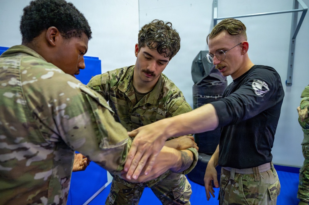 Three individuals are engaged in a close-quarters combat or self-defense training session in what appears to be a martial arts or military training facility. Two individuals are wearing military camouflage uniforms (OCP/MultiCam pattern), while a third in a black long-sleeve shirt appears to be an instructor demonstrating a wrist or arm control technique. The setting includes blue training mats and padded wall equipment consistent with a combatives training room.