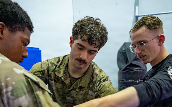 Three individuals are engaged in a close-quarters combat or self-defense training session in what appears to be a martial arts or military training facility. Two individuals are wearing military camouflage uniforms (OCP/MultiCam pattern), while a third in a black long-sleeve shirt appears to be an instructor demonstrating a wrist or arm control technique. The setting includes blue training mats and padded wall equipment consistent with a combatives training room.
