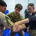 Three individuals are engaged in a close-quarters combat or self-defense training session in what appears to be a martial arts or military training facility. Two individuals are wearing military camouflage uniforms (OCP/MultiCam pattern), while a third in a black long-sleeve shirt appears to be an instructor demonstrating a wrist or arm control technique. The setting includes blue training mats and padded wall equipment consistent with a combatives training room.