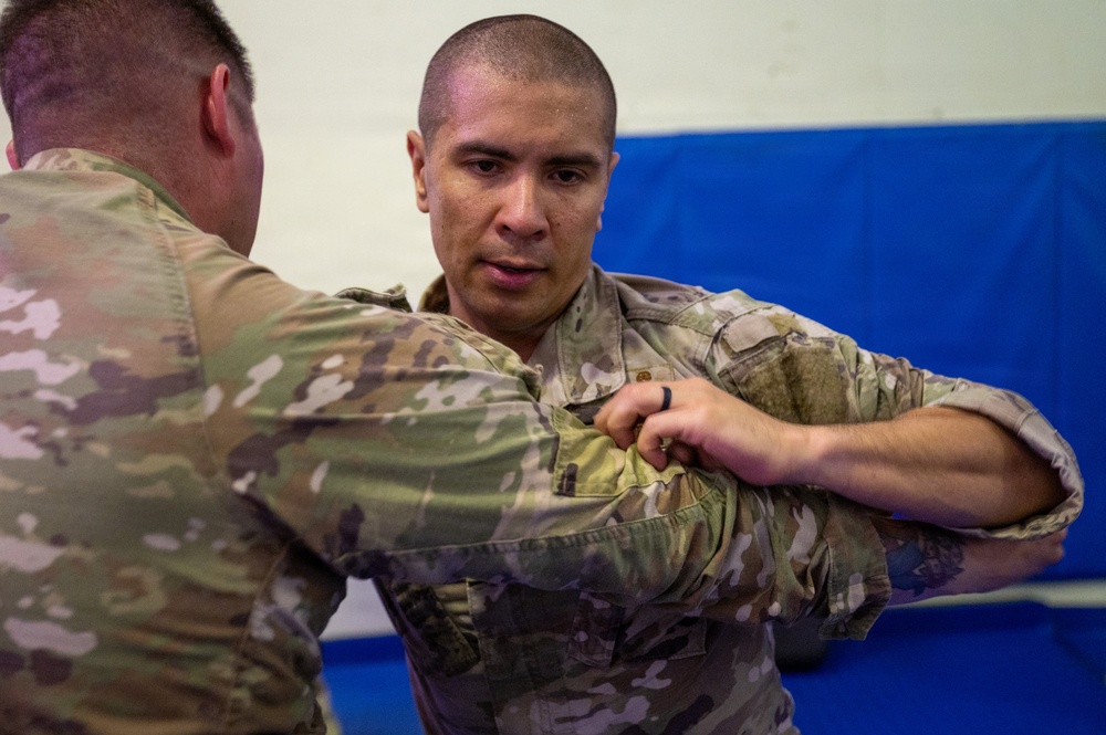 Two individuals wearing military camouflage uniforms (OCP/MultiCam pattern) are engaged in what appears to be a combatives or hand-to-hand combat training exercise. The person facing the camera is gripping the arm or uniform of the other individual in a controlled grappling technique. The setting appears to be an indoor training facility with padded blue mats visible on the wall in the background.