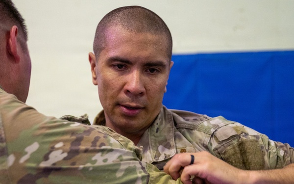 Two individuals wearing military camouflage uniforms (OCP/MultiCam pattern) are engaged in what appears to be a combatives or hand-to-hand combat training exercise. The person facing the camera is gripping the arm or uniform of the other individual in a controlled grappling technique. The setting appears to be an indoor training facility with padded blue mats visible on the wall in the background.