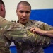 Two individuals wearing military camouflage uniforms (OCP/MultiCam pattern) are engaged in what appears to be a combatives or hand-to-hand combat training exercise. The person facing the camera is gripping the arm or uniform of the other individual in a controlled grappling technique. The setting appears to be an indoor training facility with padded blue mats visible on the wall in the background.