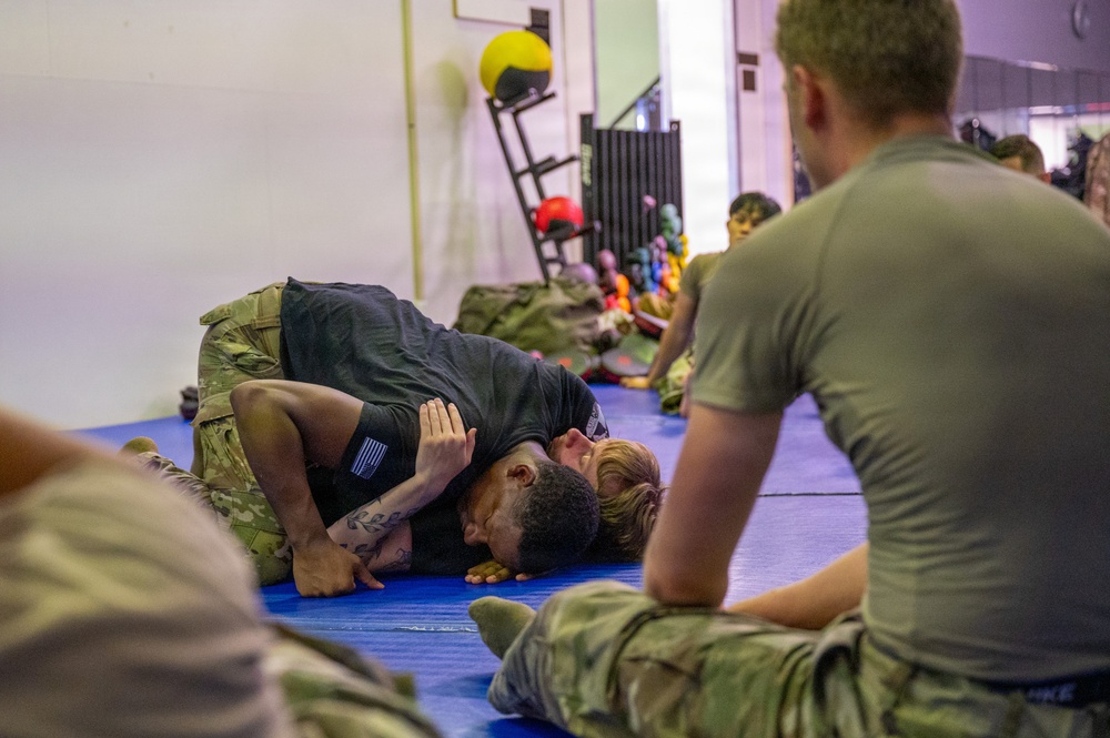 Military personnel are engaged in a combatives or ground fighting training session on blue mats inside a gymnasium or fitness facility. Two individuals are actively grappling on the mat while others in military uniforms observe from a seated position. The setting appears to be an organized hand-to-hand combat training exercise.