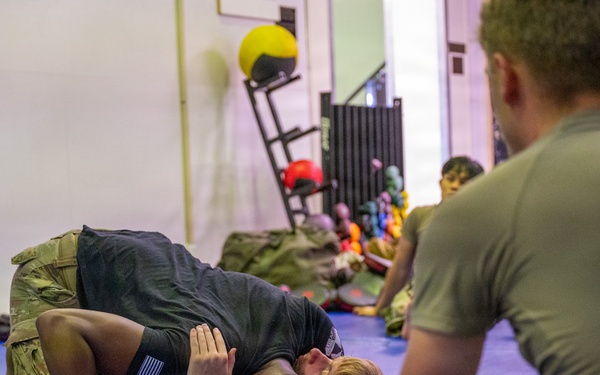 Military personnel are engaged in a combatives or ground fighting training session on blue mats inside a gymnasium or fitness facility. Two individuals are actively grappling on the mat while others in military uniforms observe from a seated position. The setting appears to be an organized hand-to-hand combat training exercise.