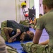 Military personnel are engaged in a combatives or ground fighting training session on blue mats inside a gymnasium or fitness facility. Two individuals are actively grappling on the mat while others in military uniforms observe from a seated position. The setting appears to be an organized hand-to-hand combat training exercise.