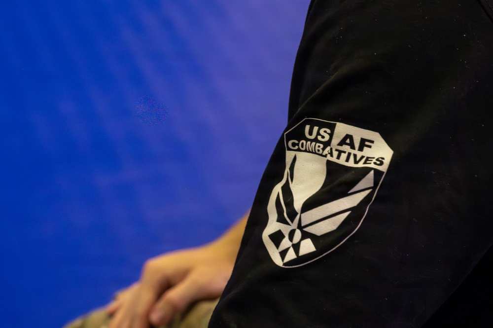 A close-up photograph of a black garment sleeve displaying a 'USAF Combatives' patch or printed logo. The insignia features the United States Air Force symbol within a shield-shaped border with the text 'US AF COMBATIVES'. The background is a solid blue surface, likely a mat or wall.