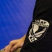 A close-up photograph of a black garment sleeve displaying a 'USAF Combatives' patch or printed logo. The insignia features the United States Air Force symbol within a shield-shaped border with the text 'US AF COMBATIVES'. The background is a solid blue surface, likely a mat or wall.