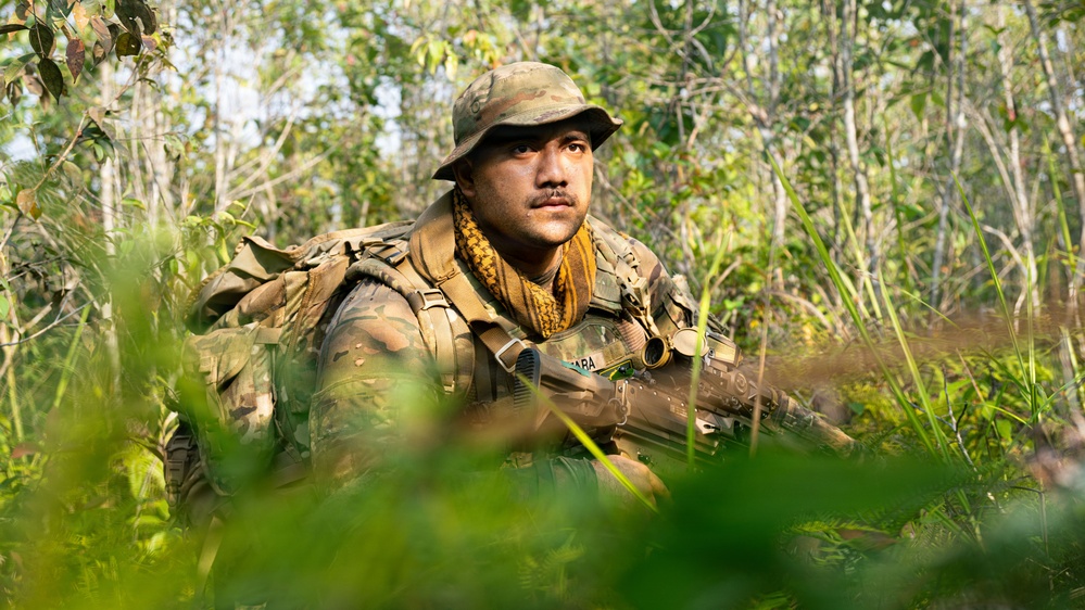 A soldier in full camouflage gear is positioned low in dense jungle vegetation, appearing to be on patrol or in a surveillance posture. He is wearing multicam/OCP-pattern camouflage uniform, a boonie hat, and a shemagh scarf, and is holding a rifle. The setting is a tropical or subtropical forest environment with lush green undergrowth.