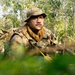 A soldier in full camouflage gear is positioned low in dense jungle vegetation, appearing to be on patrol or in a surveillance posture. He is wearing multicam/OCP-pattern camouflage uniform, a boonie hat, and a shemagh scarf, and is holding a rifle. The setting is a tropical or subtropical forest environment with lush green undergrowth.
