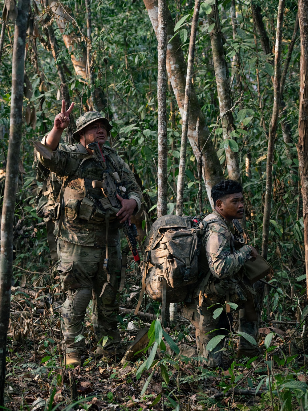 Two armed soldiers in camouflage uniforms are conducting a patrol or tactical movement through a dense tropical jungle. The lead soldier is giving a hand signal with two fingers raised, while the second soldier carries a large rucksack and appears to be in a kneeling or crouched position. Both are equipped with tactical gear consistent with a military field operation.