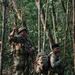Two armed soldiers in camouflage uniforms are conducting a patrol or tactical movement through a dense tropical jungle. The lead soldier is giving a hand signal with two fingers raised, while the second soldier carries a large rucksack and appears to be in a kneeling or crouched position. Both are equipped with tactical gear consistent with a military field operation.