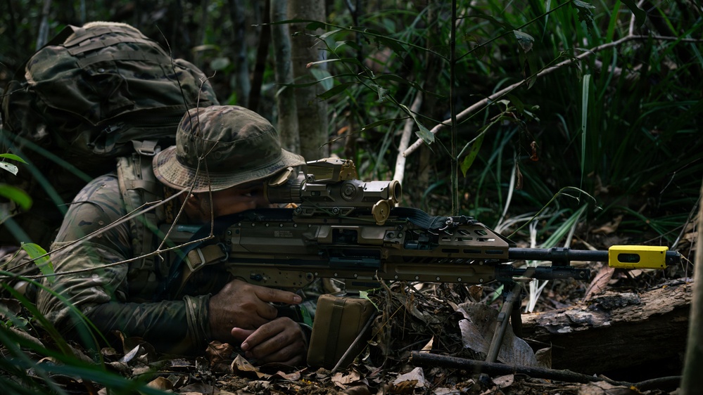 A soldier in camouflage uniform and boonie hat is prone in a jungle environment, aiming a light machine gun mounted on a bipod. The dense tropical vegetation and leaf litter suggest a jungle warfare training exercise or operation. A yellow training/safety device is visible on the muzzle end of the weapon, indicating this may be a training exercise.