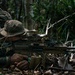 A soldier in camouflage uniform and boonie hat is prone in a jungle environment, aiming a light machine gun mounted on a bipod. The dense tropical vegetation and leaf litter suggest a jungle warfare training exercise or operation. A yellow training/safety device is visible on the muzzle end of the weapon, indicating this may be a training exercise.