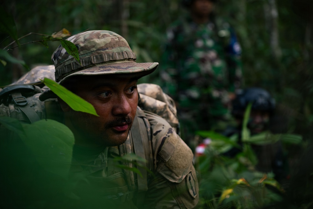 A soldier in multicam/OCP camouflage uniform and boonie hat is navigating through dense jungle vegetation while carrying a rucksack. In the background, at least two other personnel are visible wearing different camouflage patterns, suggesting a multinational military exercise or joint training operation. The setting appears to be a tropical jungle environment.