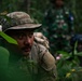 A soldier in multicam/OCP camouflage uniform and boonie hat is navigating through dense jungle vegetation while carrying a rucksack. In the background, at least two other personnel are visible wearing different camouflage patterns, suggesting a multinational military exercise or joint training operation. The setting appears to be a tropical jungle environment.
