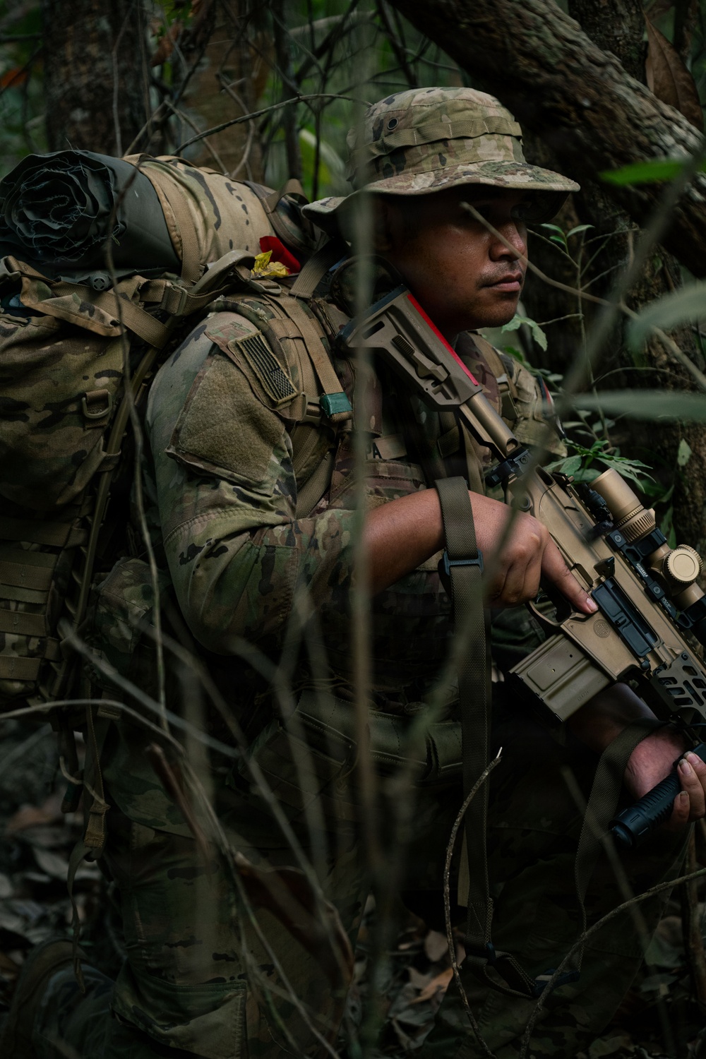 A soldier in full combat gear moves through dense jungle vegetation. The individual is equipped with a large rucksack, tactical vest, and carries a modern assault rifle with optics. The setting appears to be a jungle training or operational environment.