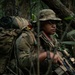 A soldier in full combat gear moves through dense jungle vegetation. The individual is equipped with a large rucksack, tactical vest, and carries a modern assault rifle with optics. The setting appears to be a jungle training or operational environment.