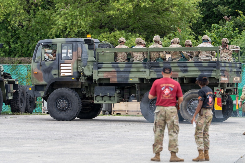 A military tactical truck (LMTV/FMTV-type) in camouflage paint is transporting a large group of soldiers in multicam/OCP uniforms in its cargo bed. Two personnel in the foreground, one wearing a red shirt with military insignia, stand observing the vehicle. The setting appears to be a military staging area or base with a paved surface and green vegetation in the background.