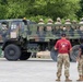 A military tactical truck (LMTV/FMTV-type) in camouflage paint is transporting a large group of soldiers in multicam/OCP uniforms in its cargo bed. Two personnel in the foreground, one wearing a red shirt with military insignia, stand observing the vehicle. The setting appears to be a military staging area or base with a paved surface and green vegetation in the background.