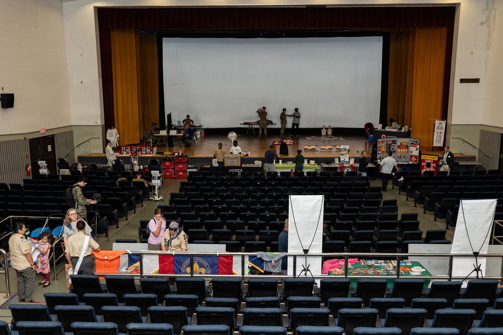 An auditorium or theater venue is being used for what appears to be a community resource fair or military family support event. Vendor and information tables are set up along the front of the stage and along the back row of seating, with participants in various uniforms including military and Boy Scouts visible. The main seating area remains largely empty while organizers and attendees interact at the booths.