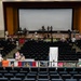 An auditorium or theater venue is being used for what appears to be a community resource fair or military family support event. Vendor and information tables are set up along the front of the stage and along the back row of seating, with participants in various uniforms including military and Boy Scouts visible. The main seating area remains largely empty while organizers and attendees interact at the booths.