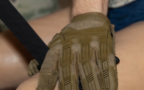 A gloved hand wearing a tan/olive tactical Mechanix-brand glove is applying or adjusting a tourniquet on a person's forearm or lower limb. A black strap tourniquet with a windlass rod is visible, consistent with a CAT (Combat Application Tourniquet) or similar device. A bruise or wound mark is visible on the limb, suggesting this is either a medical training exercise or an actual first-aid application.