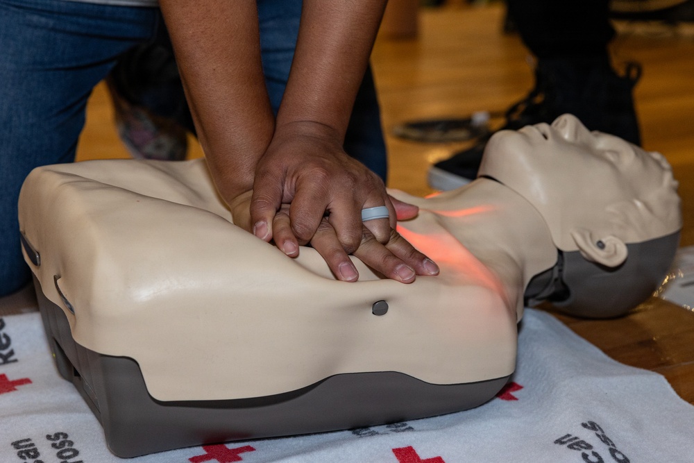 A person is performing CPR chest compressions on a training manikin during what appears to be a first aid or CPR certification course. The manikin features a red feedback light indicating compression depth or placement accuracy. A Red Cross branded mat is visible beneath the manikin, confirming an organized training context.