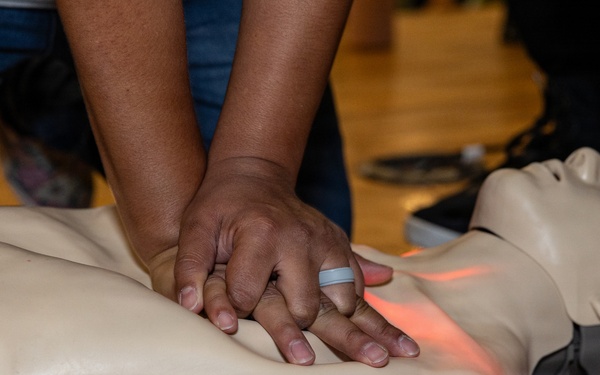A person is performing CPR chest compressions on a training manikin during what appears to be a first aid or CPR certification course. The manikin features a red feedback light indicating compression depth or placement accuracy. A Red Cross branded mat is visible beneath the manikin, confirming an organized training context.