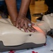 A person is performing CPR chest compressions on a training manikin during what appears to be a first aid or CPR certification course. The manikin features a red feedback light indicating compression depth or placement accuracy. A Red Cross branded mat is visible beneath the manikin, confirming an organized training context.