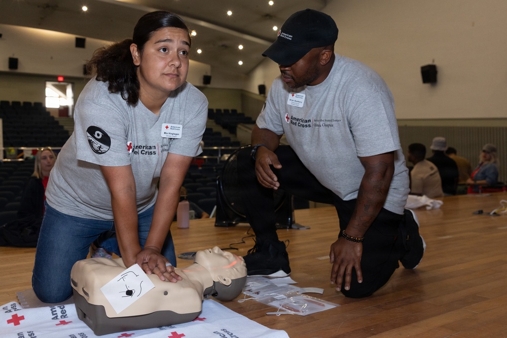 Two American Red Cross volunteers in matching gray shirts are conducting a CPR training session on a wooden gymnasium floor. A woman is performing chest compressions on a CPR training mannequin while a man observes and supervises her technique. The setting appears to be a large auditorium or gymnasium with additional trainees visible in the background.