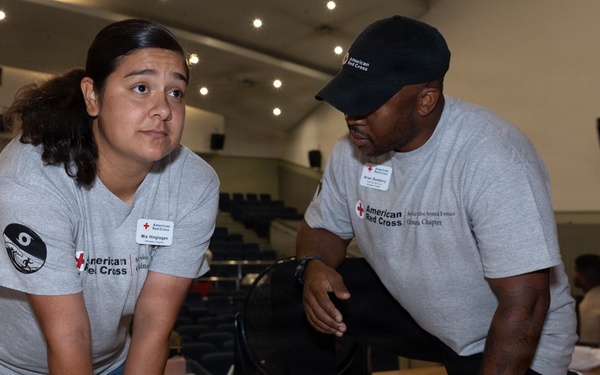 Two American Red Cross volunteers in matching gray shirts are conducting a CPR training session on a wooden gymnasium floor. A woman is performing chest compressions on a CPR training mannequin while a man observes and supervises her technique. The setting appears to be a large auditorium or gymnasium with additional trainees visible in the background.