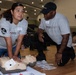 Two American Red Cross volunteers in matching gray shirts are conducting a CPR training session on a wooden gymnasium floor. A woman is performing chest compressions on a CPR training mannequin while a man observes and supervises her technique. The setting appears to be a large auditorium or gymnasium with additional trainees visible in the background.