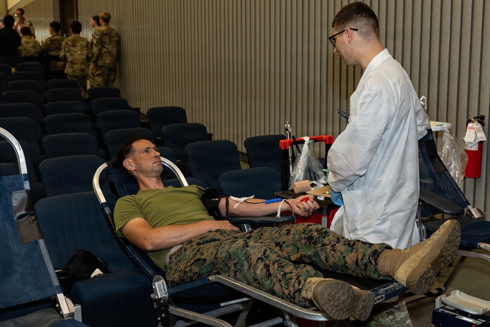 A military service member in camouflage uniform and olive green t-shirt is donating blood while reclined on a donor chair. A medical technician in a white lab coat is attending to the donor, managing blood collection equipment. The setting appears to be a blood drive held in a large auditorium or assembly hall, with other military personnel visible in the background.