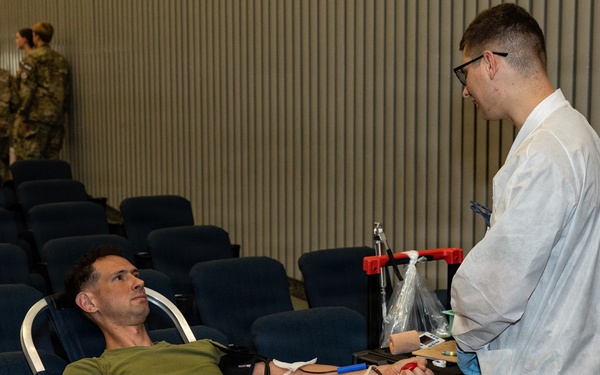 A military service member in camouflage uniform and olive green t-shirt is donating blood while reclined on a donor chair. A medical technician in a white lab coat is attending to the donor, managing blood collection equipment. The setting appears to be a blood drive held in a large auditorium or assembly hall, with other military personnel visible in the background.