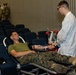 A military service member in camouflage uniform and olive green t-shirt is donating blood while reclined on a donor chair. A medical technician in a white lab coat is attending to the donor, managing blood collection equipment. The setting appears to be a blood drive held in a large auditorium or assembly hall, with other military personnel visible in the background.