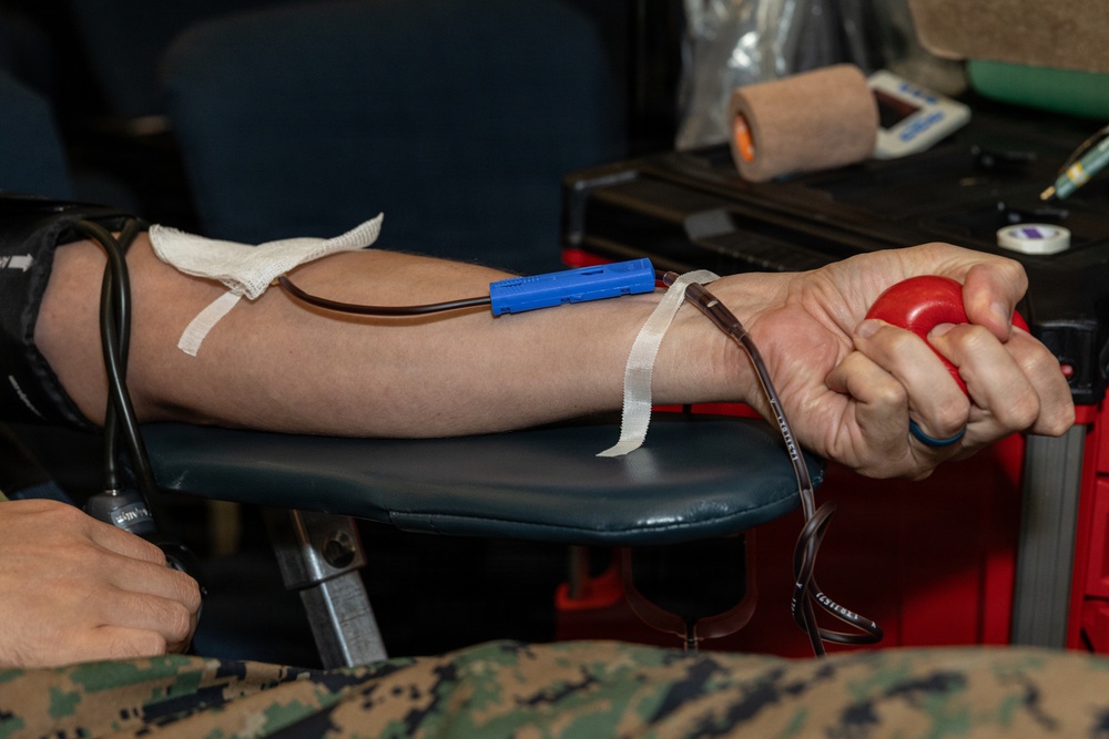 The image shows a person donating blood, with a needle and tubing inserted into the inner arm secured with medical tape. The donor is squeezing a red stress ball to help stimulate blood flow during the donation process. Medical equipment and supplies are visible in the background on a table.