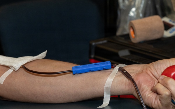 The image shows a person donating blood, with a needle and tubing inserted into the inner arm secured with medical tape. The donor is squeezing a red stress ball to help stimulate blood flow during the donation process. Medical equipment and supplies are visible in the background on a table.