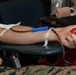 The image shows a person donating blood, with a needle and tubing inserted into the inner arm secured with medical tape. The donor is squeezing a red stress ball to help stimulate blood flow during the donation process. Medical equipment and supplies are visible in the background on a table.