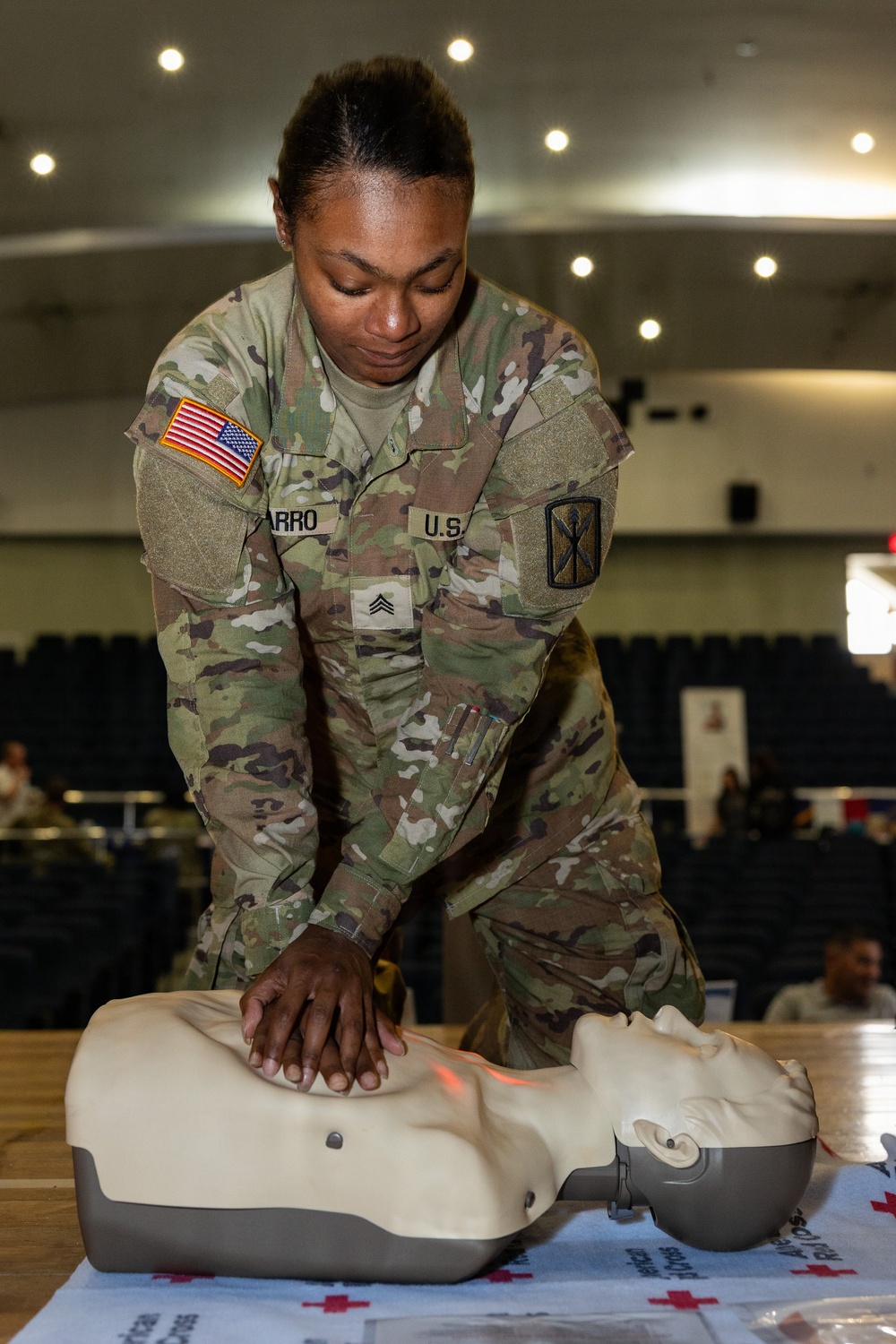A U.S. Army Sergeant in OCP (Operational Camouflage Pattern) uniform is performing chest compressions on a CPR training mannequin. The soldier, whose name tape partially reads 'ARRO' and wears a Sergeant rank insignia, is demonstrating proper CPR technique in what appears to be an indoor training facility or auditorium. A red-light indicator on the mannequin suggests it is an electronic feedback training device.