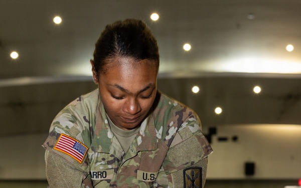 A U.S. Army Sergeant in OCP (Operational Camouflage Pattern) uniform is performing chest compressions on a CPR training mannequin. The soldier, whose name tape partially reads 'ARRO' and wears a Sergeant rank insignia, is demonstrating proper CPR technique in what appears to be an indoor training facility or auditorium. A red-light indicator on the mannequin suggests it is an electronic feedback training device.