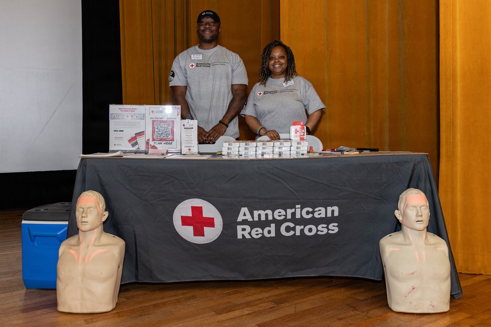 Two American Red Cross volunteers stand behind a branded table at what appears to be a community outreach or health fair event. The table displays informational materials, QR codes, and small packaged items, while two CPR training mannequin torsos are positioned in front of the table. Both individuals are wearing matching gray American Red Cross t-shirts and appear engaged and welcoming.