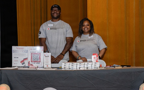 Two American Red Cross volunteers stand behind a branded table at what appears to be a community outreach or health fair event. The table displays informational materials, QR codes, and small packaged items, while two CPR training mannequin torsos are positioned in front of the table. Both individuals are wearing matching gray American Red Cross t-shirts and appear engaged and welcoming.