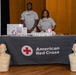 Two American Red Cross volunteers stand behind a branded table at what appears to be a community outreach or health fair event. The table displays informational materials, QR codes, and small packaged items, while two CPR training mannequin torsos are positioned in front of the table. Both individuals are wearing matching gray American Red Cross t-shirts and appear engaged and welcoming.