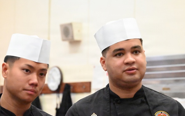 Two United States Marine Corps personnel are wearing black chef uniforms with USMC insignia patches and white chef toques, standing in what appears to be a military kitchen facility. The individual on the right has a name tape reading 'LANCELOT' and displays a corporal rank insignia. Both individuals appear to be attentive, possibly receiving instructions or observing a demonstration.