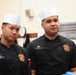 Two United States Marine Corps personnel are wearing black chef uniforms with USMC insignia patches and white chef toques, standing in what appears to be a military kitchen facility. The individual on the right has a name tape reading 'LANCELOT' and displays a corporal rank insignia. Both individuals appear to be attentive, possibly receiving instructions or observing a demonstration.