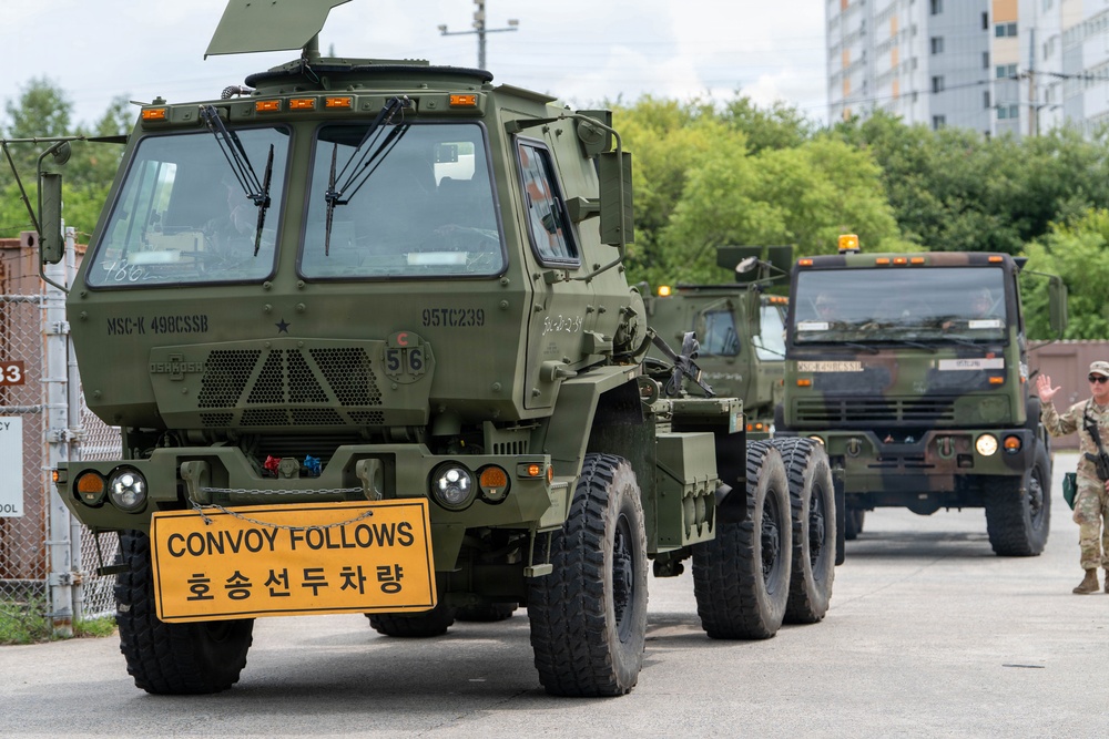 A military convoy of olive-green heavy tactical trucks is moving through what appears to be a U.S. military installation in South Korea. The lead vehicle displays a bilingual 'CONVOY FOLLOWS / 호송선두차량' sign in English and Korean, indicating this is the front vehicle of a convoy. A soldier in uniform is visible on the right side, apparently directing or observing the convoy movement.