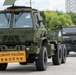 A military convoy of olive-green heavy tactical trucks is moving through what appears to be a U.S. military installation in South Korea. The lead vehicle displays a bilingual 'CONVOY FOLLOWS / 호송선두차량' sign in English and Korean, indicating this is the front vehicle of a convoy. A soldier in uniform is visible on the right side, apparently directing or observing the convoy movement.