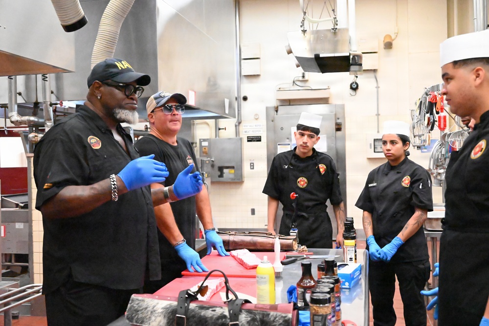 A culinary instruction session is taking place in a professional kitchen, where two instructors wearing black shirts and blue latex gloves appear to be demonstrating cooking techniques to three students. The students are dressed in black chef uniforms with matching patches/logos, and one wears a traditional white chef's toque. A red cutting board with meat and various condiment bottles and sauces are visible on the work table.
